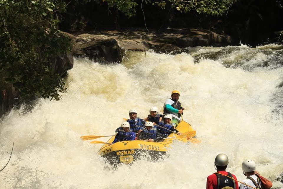 Bote encarando corredeira forte cercada de mata, com equipe de apoio na margem